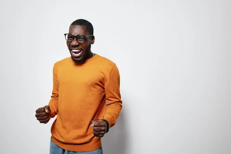 African American man with dark skin wearing glasses and orange sweater smiling and celebrating with clenched fists on white background. Happy male portrait with casual style.