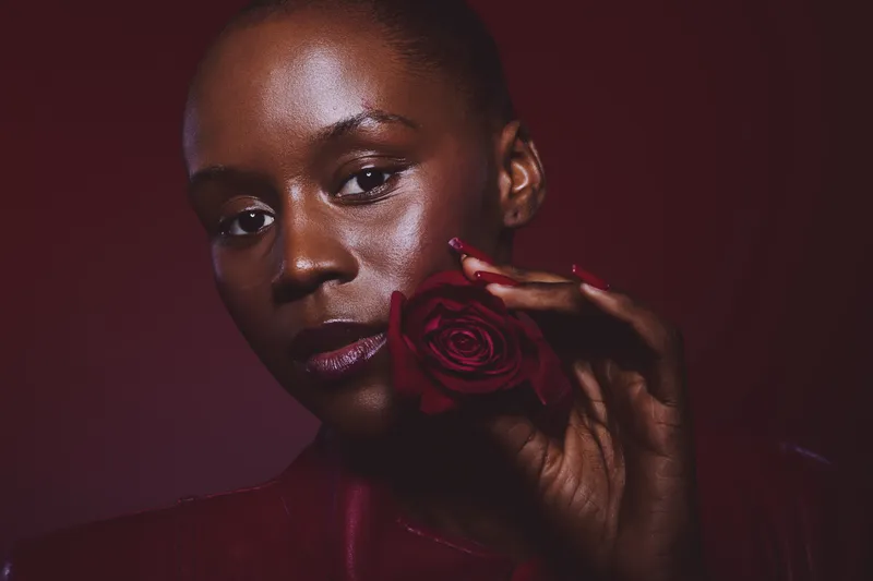 Close-up portrait of a dark skinned African woman holding a red rose near her face. The image features dramatic lighting and a deep red background, highlighting beauty and elegance.