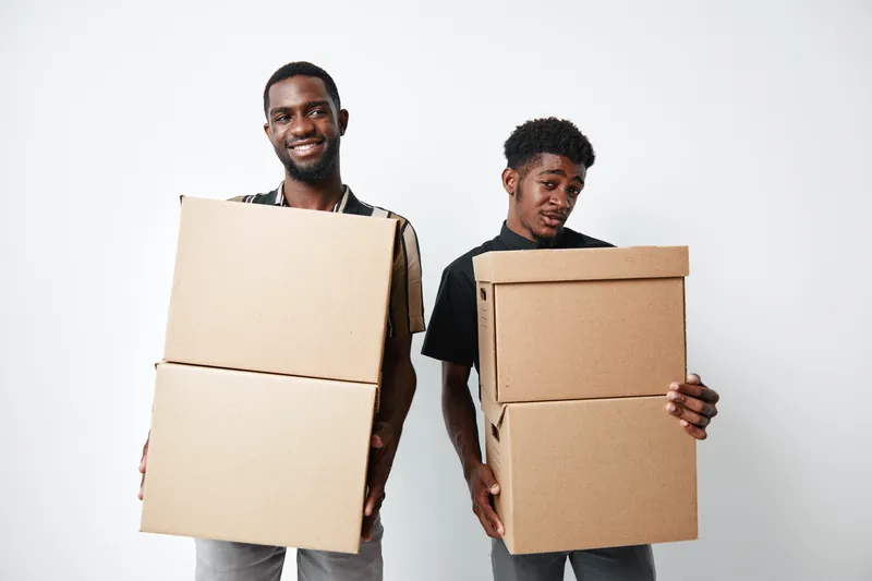 Two young black men holding cardboard boxes for moving or delivery on white background. African ethnicity, casual clothing, teamwork and logistics concept with smiling and focused expressions.