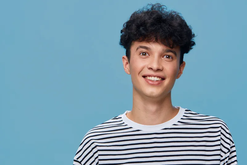 smiling young man with curly hair wearing striped shirt against blue background, cheerful expression, casual style, happy mood, studio portrait, light skin tone