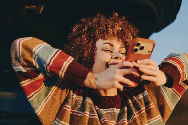 Woman lifestyle portrait, curly hair, striped sweater, smiling while holding phone and using smartphone, cozy indoor background, warm light, relaxed casual everyday moment, copy space