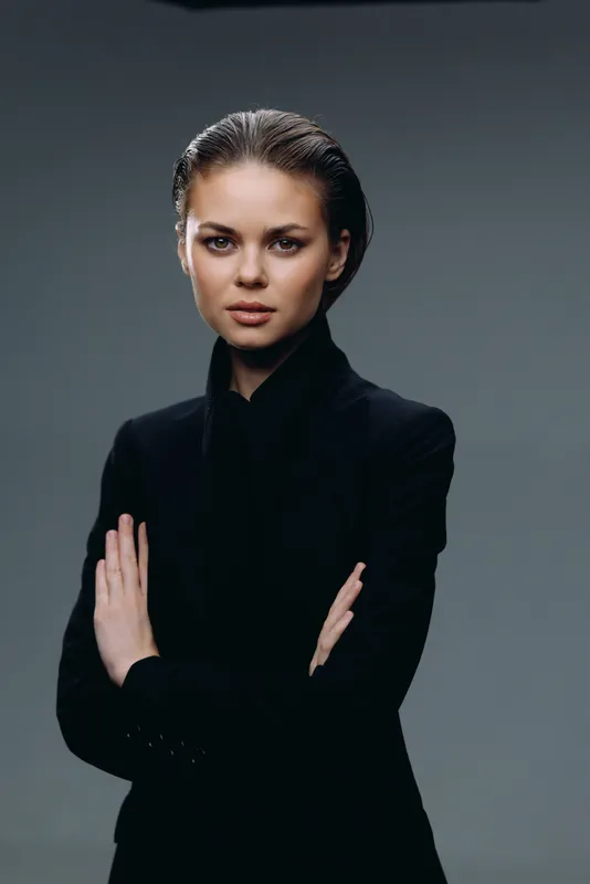 Confident woman wearing black jacket poses with crossed arms against dark gray background, displaying strong expression and professional style.