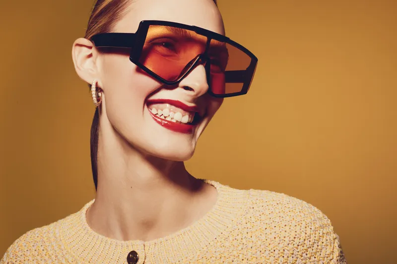 Smiling woman with large sunglasses and red lipstick wears yellow knitted sweater. Studio portrait on warm brown background. Concept of fashion, style, beauty, and confident mood.