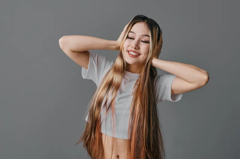 Young woman with long hair wearing white t shirt holds hands on head with eyes closed smiling against grey background. Beauty, happiness and relaxation concept.