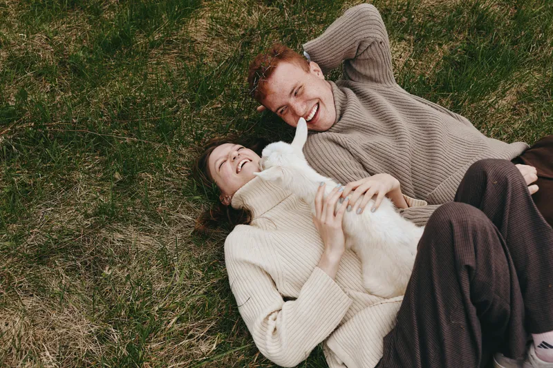 Cheerful couple outdoors, lying on grass with a gentle goat kid, relaxed pose, warm sweater textures, sunlit moment, intimate connection, natural colors, rustic meadow backdrop