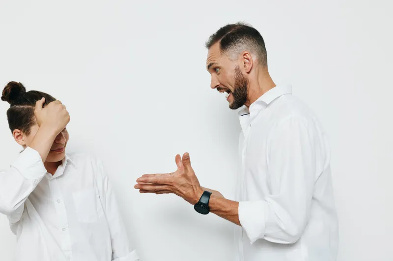 A professional businessman stands against a vibrant colored background, displaying strong emotion as he gestures toward a colleague with open hands.