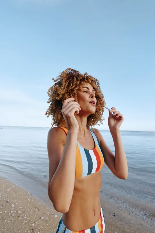 Woman on beach, holding a shell near her ear, curly hair in motion, wearing a colorful bikini, smiling by the sea, summer vacation mood, under clear sky, gentle breeze, waves, background
