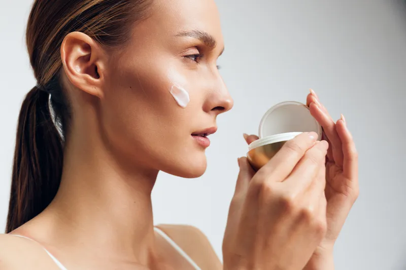 Young woman applies face cream on cheek holding cosmetic jar close to face with focused expression isolated on light background in skincare beauty routine concept.