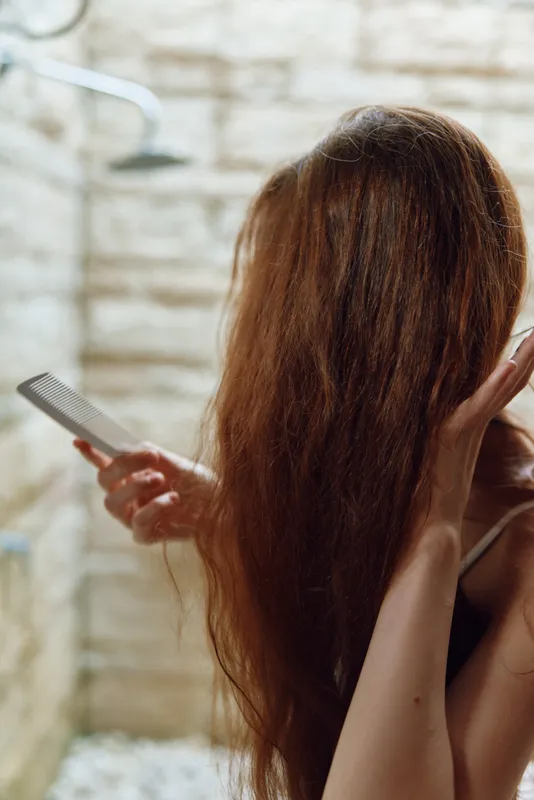 woman washing long brown hair under outdoor shower with stone wall background in natural light, showing self-care and relaxation in a serene setting