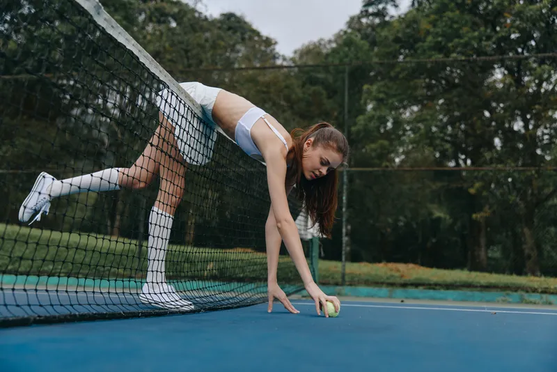 Young athletic woman in white sportswear reaching for a tennis ball on a blue court, showcasing determination and focus amidst a green outdoor background