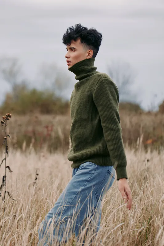 Young man in a knit turtleneck walks through tall dry grass, captured in a candid portrait emphasizing natural authenticity and subtle credibility, a raw outdoor moment with honest emotion.