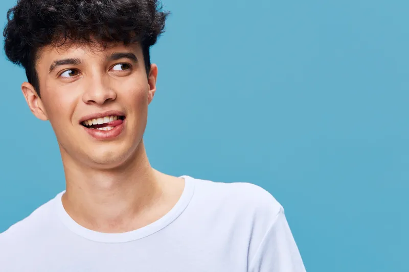 young man with curly hair smiling playfully wearing white t-shirt isolated on blue background, casual style and youthful expression, vibrant colors