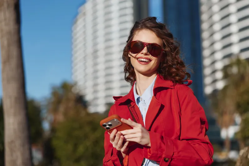 Cheerful woman outdoors wearing red coat, sunglasses, and blue shirt, smiling while using a smartphone in a sunny urban park setting with natural light and a vibrant background