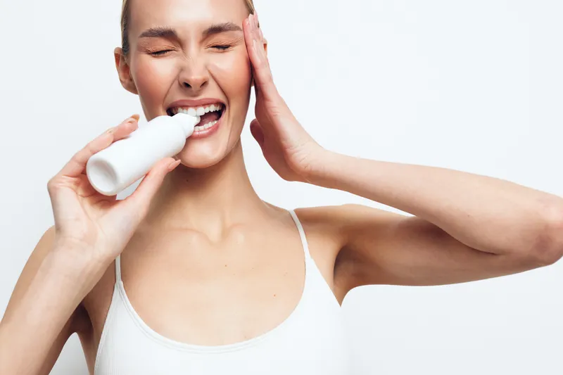 Young woman with brunette hair in a white tank top smiles while applying foam from a cosmetic device to her cheek, isolated on a light background.