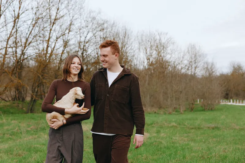 Couple walking in a grassy park smiling while holding a small dog, casual outfits, candid lifestyle moment, outdoor companionship, warm mood, fresh air, natural light, tranquil countryside scene.