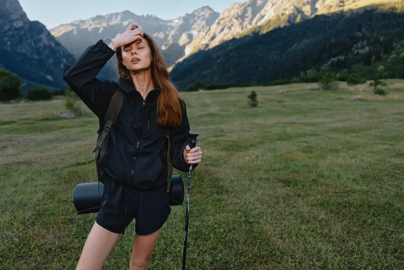 A cheerful hiker stands in a wide mountain meadow, adjusting her hair and smiling, with a camera and backpack, radiating energy, optimism, and outdoor adventure.