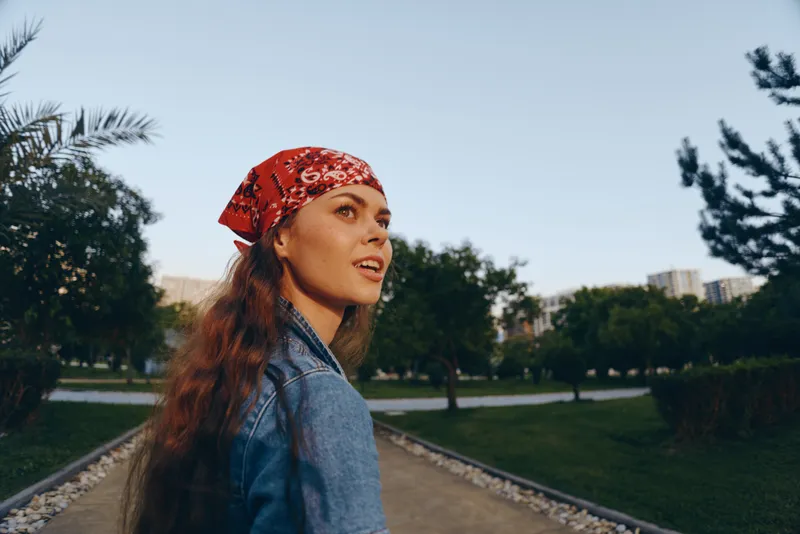 young woman with red bandana and long hair wearing denim jacket outdoors in park with green trees in sunny weather during daytime
