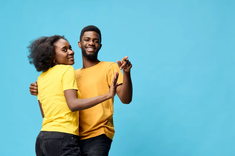 Young African couple dancing joyfully together in vibrant yellow outfits against a light blue background, expressing happiness and connection
