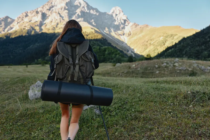 A lone hiker with a backpack and a rolled mat stands in a vast alpine meadow, gazing toward rugged peaks under clear skies, capturing a sense of bold adventure.