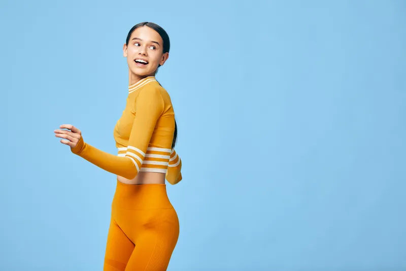 Vibrant fitness portrait of a woman in orange athletic wear posing mid movement against a blue backdrop, showcasing sport styling, energy and confidence.