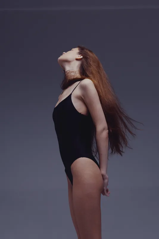 slender woman in black bodysuit posing with head tilted back against gray background, minimalistic fashion portrait