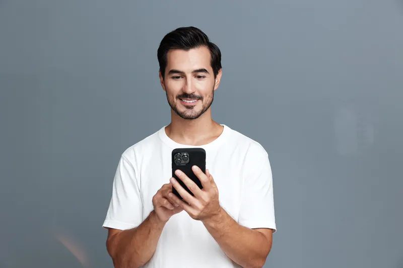 Smiling man holding smartphone, dressed in casual white t-shirt, looking at screen with friendly expression against gray background. Modern lifestyle concept.