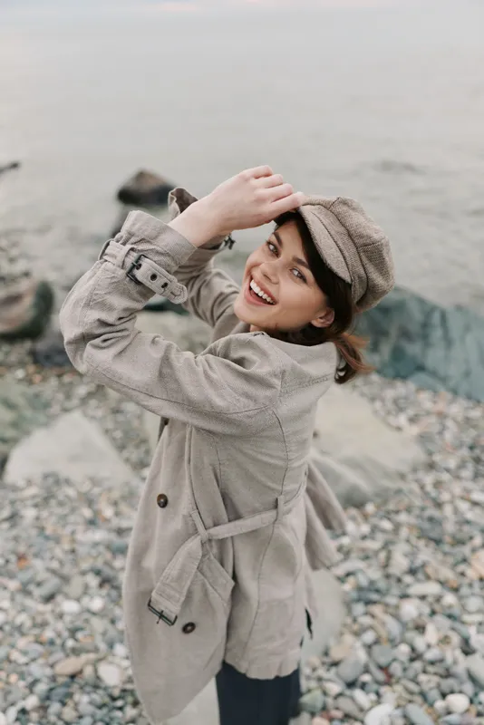 Smiling woman in beige coat and hat poses on rocky beach near water. Natural light highlights joyful mood and casual fashion style in outdoor coastal environment.