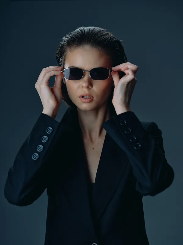 Young woman wearing sunglasses and a black blazer with a confident expression on dark background