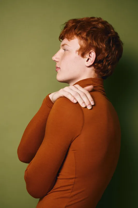 Profile portrait of young man with red hair wearing brown turtleneck standing against green background with folded arms and thoughtful expression in studio lighting.