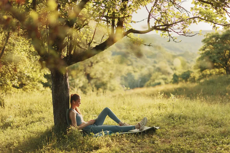 Young woman enjoying a sunny day in a serene green park, reading a book under a tree, showcasing relaxation and connection with nature