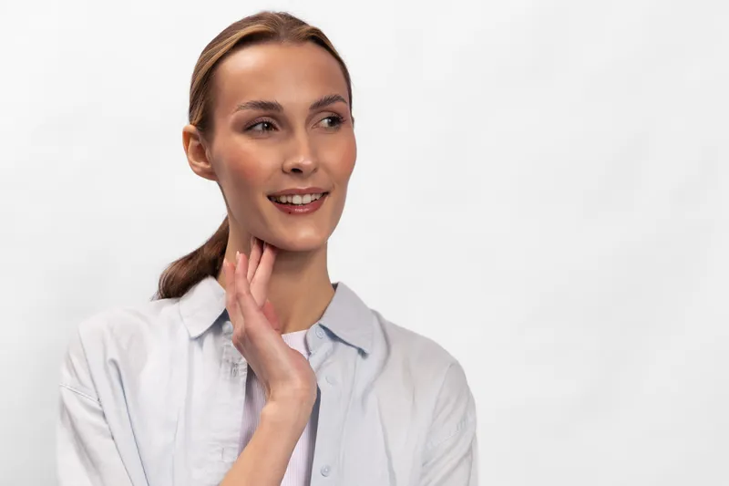 Young woman with clear skin and light brown hair smiles gently wearing a white shirt against white background. Concept for beauty, health, natural look, and lifestyle portrait.