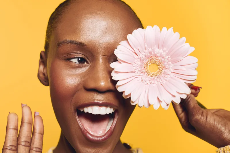 Happy African woman with dark skin holds pink flower near face and smiles widely. Yellow background creates bright, cheerful mood. Concept of natural beauty, joy, and floral freshness.
