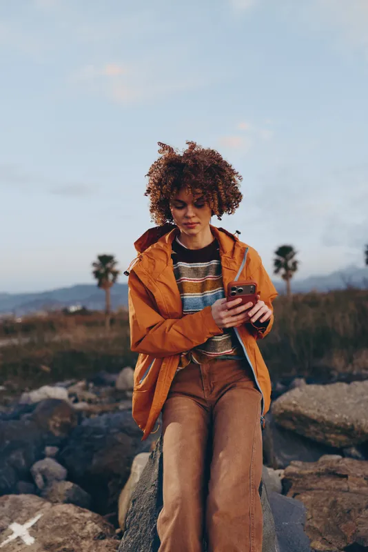 Woman lifestyle outdoors mountain landscape, standing on rocks, holding smartphone, wearing coat, curly hair, relaxed walking moment, scenic horizon under soft sky