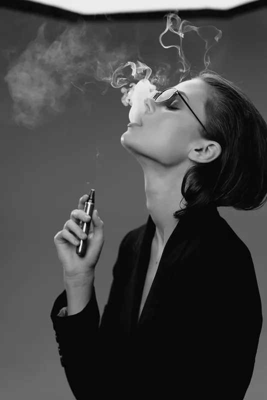 Black and white portrait of a woman vaping and blowing smoke in a dark background with glasses and stylish hair.