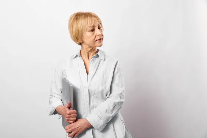 Elderly woman holding a folder and book in a studio portrait, white background, calm serious expression, soft light, standing pose, copy space, looking to the side quietly