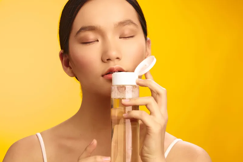 asian woman with closed eyes holds a skincare bottle near her face, standing against a bright yellow background, conveying calm, self care, and a serene beauty ritual.