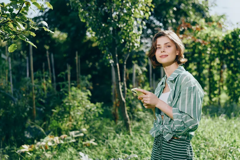 A young woman stands in a sunlit garden, gazing at her phone while surrounded by lush greenery and blooming plants.
