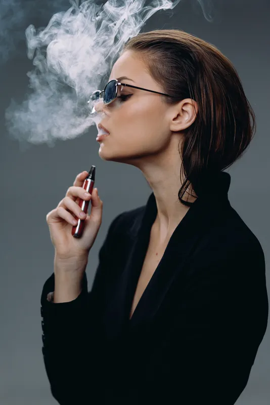 Stylish woman vaping with smoke exhaling, wearing sunglasses in a dark outfit, profile portrait with modern and edgy vibe
