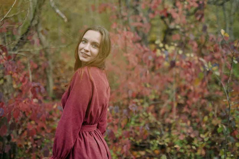 A candid portrait of a young woman in a simple red dress standing amid autumn foliage, conveying an authentic, natural moment with soft light and a genuine, warm expression.