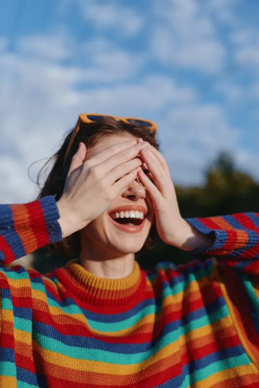Woman smiling wearing a colorful striped sweater, hands covering eyes playfully, sunglasses on head, casual young adult outdoors under blue sky, joyful lifestyle portrait with warm expression.