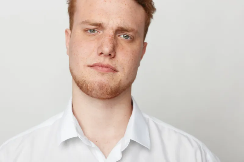 Close-up portrait of serious young man with red hair and freckles wearing white shirt against plain background. Concept of confidence, youth, and natural appearance.
