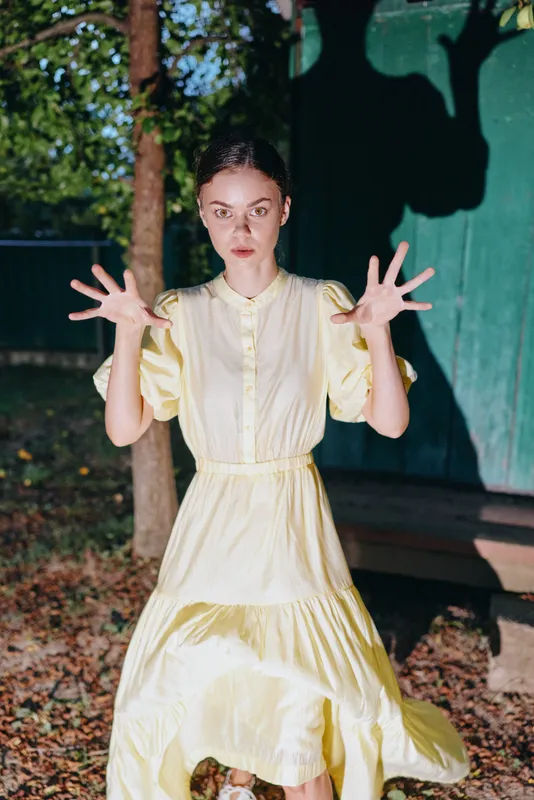 Woman in a flowing yellow dress poses dynamically outdoors, palms raised and focused expression, sunlight casting dramatic shadows on a green backdrop and ground