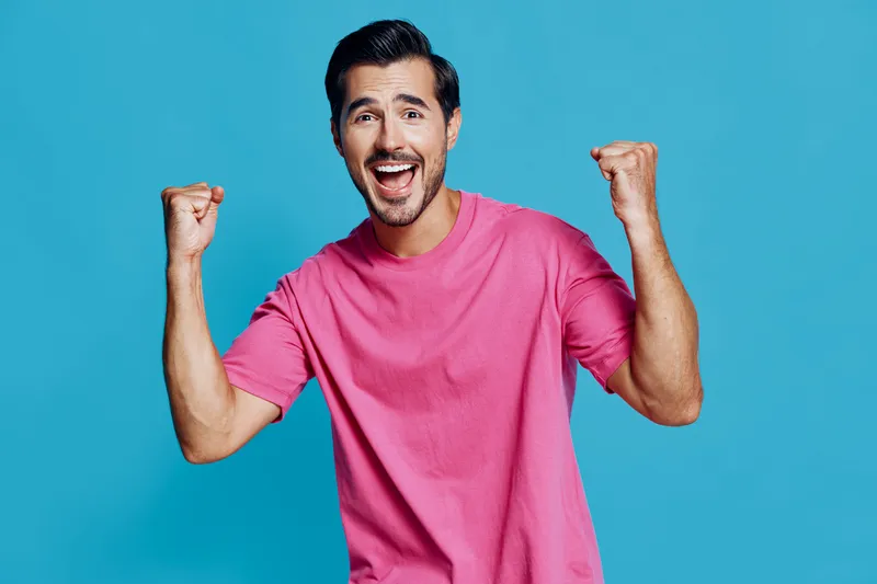 Excited cheerful young caucasian man wearing pink t-shirt raises fists in celebration showing joy and happiness isolated on bright blue background. People lifestyle concept