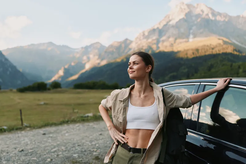 Young woman stands beside a car in a rugged mountain landscape, posing with confidence after a roadside pause