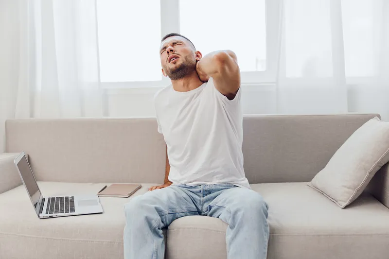 Painful man stretching his neck while sitting on a modern couch. The image captures discomfort, home office environment, and casual attire for a relatable vibe.