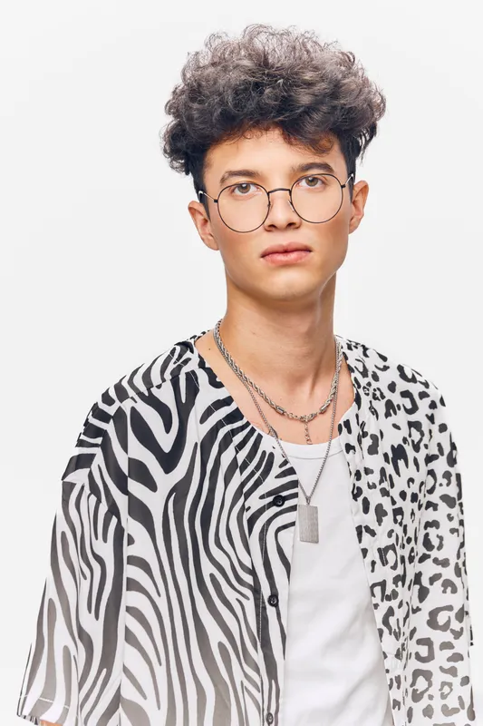 young man with curly hair wearing glasses and a zebra print shirt on white background, looking confident with layered silver necklaces and casual style