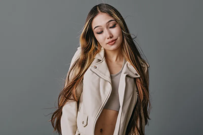 Young woman with long hair wearing casual beige jacket and tank top posing with eyes closed on gray background. Fashion model with natural expression and relaxed posture.