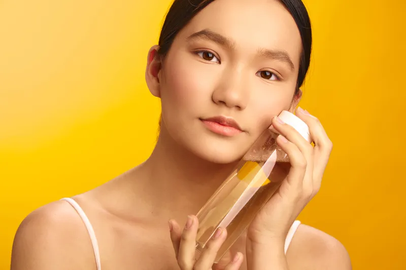 Asian woman is photographed in a bright studio, focusing on a skincare bottle as she applies product with calm confidence, creating a clean, modern aesthetic.