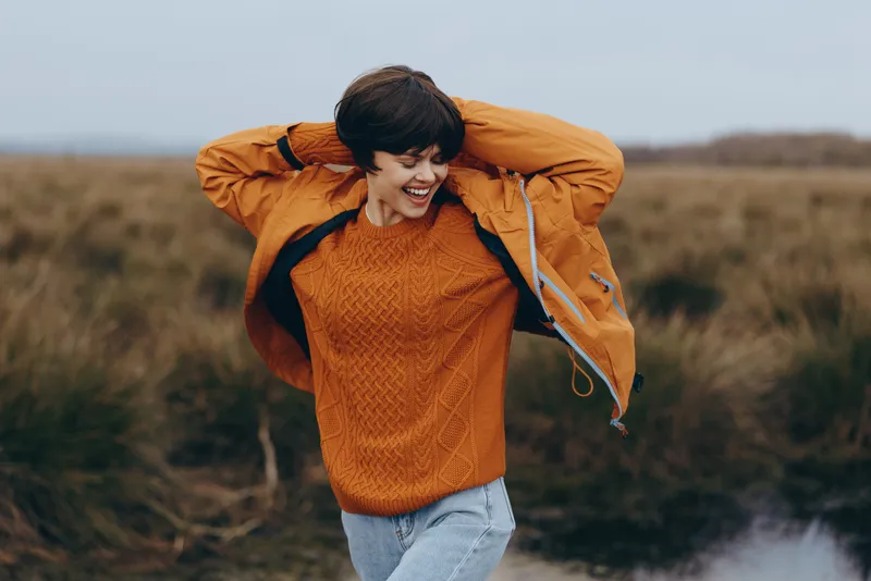 Woman lifestyle outdoors field autumn sweater jacket smiling, she lifts her jacket above head, orange sweater and blue denim jeans, wind moves her hair, she walks on a countryside path, soft