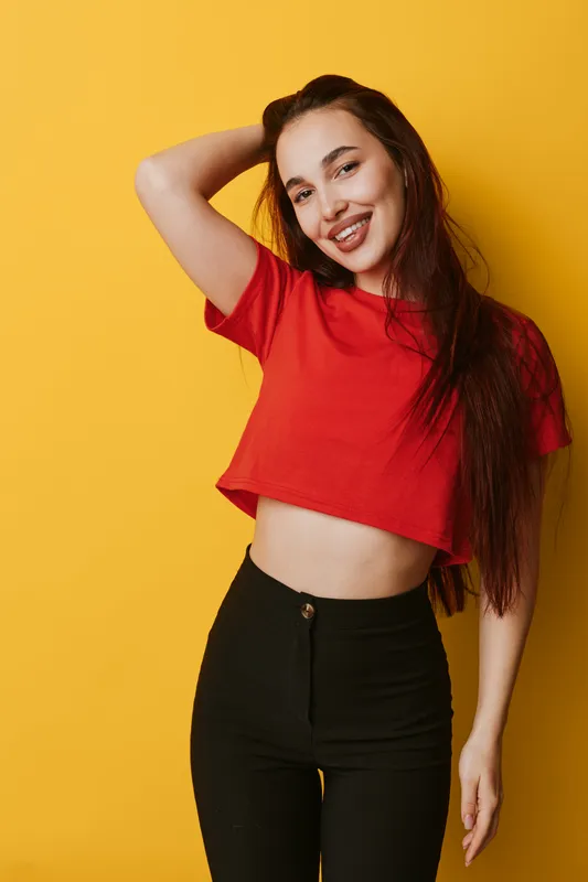 Young woman smiling and posing with hand in hair wearing red crop top and black pants standing against yellow background in casual style and confident mood.